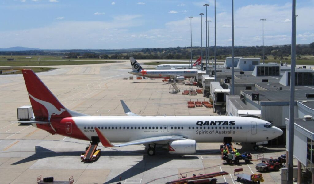 Qantas aircraft at airport gate with ground crew and luggage carts, Jetstar and other planes visible in background.